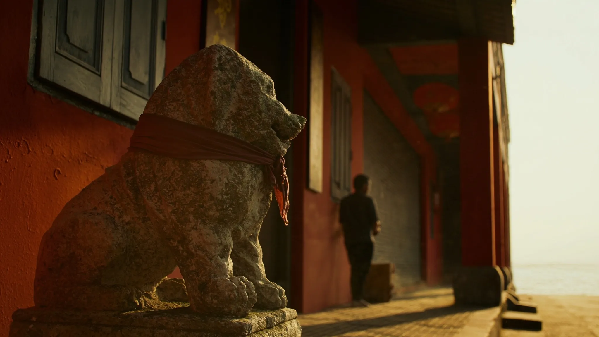 A weathered stone dog draped in faded red cloth keeps watch over a terracotta shophouse five-foot-way at golden hour, a lone figure walking toward the sea at the street's end.