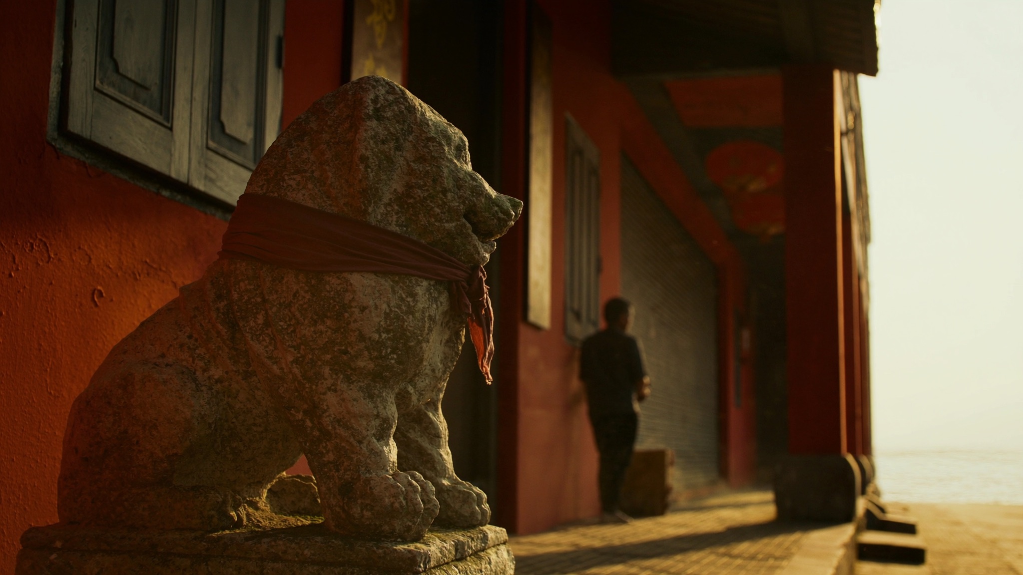 A weathered stone dog draped in faded red cloth keeps watch over a terracotta shophouse five-foot-way at golden hour, a lone figure walking toward the sea at the street's end.